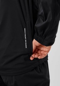 Main tirant la fermeture éclair sur la poche d'une veste noire avec le texte « SWEDMOUNT ON COURSE » sur le côté de la poche, portant un pantalon noir assorti.