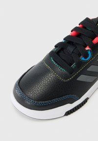 Des baskets synthétiques noires présentent une surface texturée, des coutures colorées en bleu et jaune, et une semelle en caoutchouc blanc avec des motifs de traction.