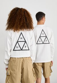 Hvite langermede skjorter med svart trekantet design som har bokstavene H, U og F på ryggen, kombinert med beige cargo-shorts.