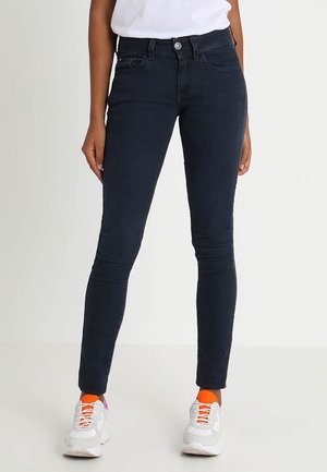 Kobieta w ciemnoniebieskich obcisłych dżinsach, białych trampkach z pomarańczowymi skarpetkami i białej bluzce, stojąca na jednolitym jasnym tle.