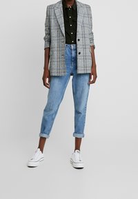 Grå rutigt kavaj med två knappar, svart skjorta, uppvikta blå jeans och vita sneakers. Kavajen har en strukturerad design.