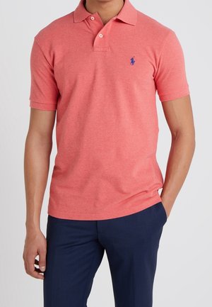 Mann trägt ein korallenfarbenes kurzärmeliges Polo mit zwei Knöpfen und dunkelblaue Hose, steht vor einem schlichten hellen Hintergrund.