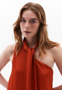 Rotes ärmelloses Oberteil mit hohem Kragen und geknotetem Akzent. Glatter Stoff, minimalistisches Design und eine Schulter freigelegt.