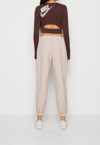 Brun långärmad croptop med utskuren rygg och vit Nike-logotyp; ljusbeiga, lätta, elastiska byxor med sidofickor.