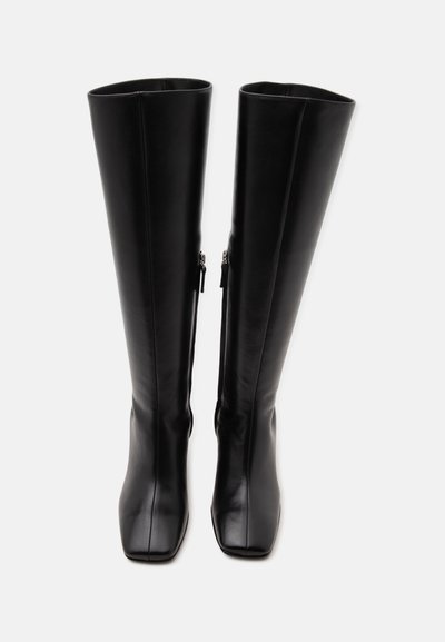 Paire de bottes hautes en cuir noir avec bouts carrés et fermetures éclair latérales, debout sur un fond blanc uni.
