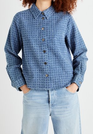 Kvinde iført en tekstureret blå skjorte med knapper og lange ærmer, let stoppet ned i lyseblå jeans, hænderne i lommerne.