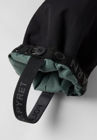 Zwarte en groene waterdichte jasmanchet met een gathered elastische opening, met een knop en labeldetails op zwarte stof.