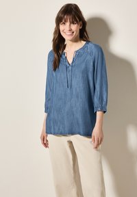 Top in chambray azzurro con scollatura a V, polsini elasticizzati e cordoncino. Abbinalo a pantaloni beige a gamba larga. Tessuto morbido, vestibilità rilassata.