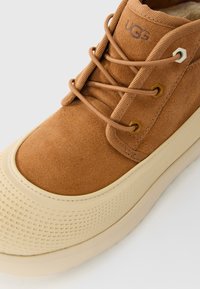 Boot UGG en daim marron avec une semelle en caoutchouc beige ronde et des lacets marron assortis, montrant un gros plan de l'orteil et de la zone de laçage.