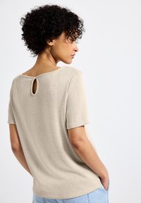 Beige kortärmad topp med en texturerad yta, med en nyckelhålsdetalj på baksidan och en avslappnad passform. Ljusblå jeans synliga.