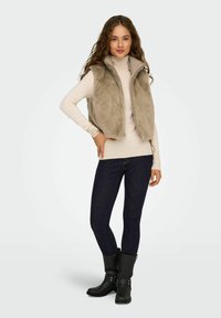Beige nepbont vest met hoge kraag, ritsluiting, gecombineerd met een crèmekleurige coltrui en donkerdenim skinny jeans. Zwarte enkellaarsjes.