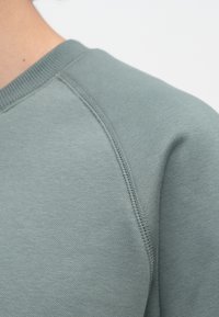Šviesiai žalias džemperis su siūta iškirpte ir tekstūruota medžiaga, iš arti matomas pečių siūlių detalizavimas ir siuvimas.