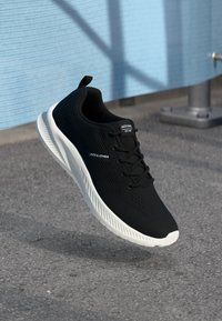 Sorte atletiske sko med en tekstureret overdel, hvid sål og logo på tungen, der har seks snørehuller og en pull-tab bagpå.