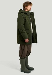 Grön vattentät parka med avtagbar huva, sidofickor och tryckknappar, kombinerad med bruna friluftsbysshorts och gröna gummistövlar.