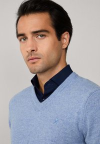 Světle modrý svetr s výstřihem do V, vyrobený z měkkého materiálu, s malým modrým vyšitým logem na hrudi, nesený přes tmavě modrou košili s límečkem.