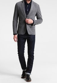 Harmaa bleiseri, jossa on lovihalkioiset kaulukset, tummanruudullisen paidan päällä, yhdistettynä tummiin farkkuihin ja mustiin kenkiin. Rentoa mutta istuvaa tyyliä.