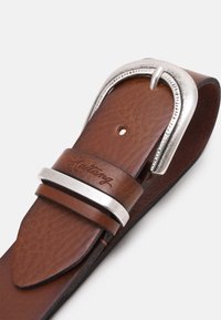 Brauner Leder-Gürtel mit einer silberfarbenen Metallschnalle, die eine strukturierte Oberfläche und eine gravierte Markeninschrift am Riemen aufweist.