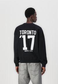 Hanorac negru cu detalii albe cu text "TORONTO '17 MAPLE LEAFS," având guler rotund și mansete ribbate. Purtați cu blugi gri.