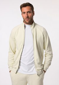 Chaqueta de cremallera color crema claro hecha de tejido suave, con cuello alto y puños acanalados, combinada con una capa base blanca debajo.
