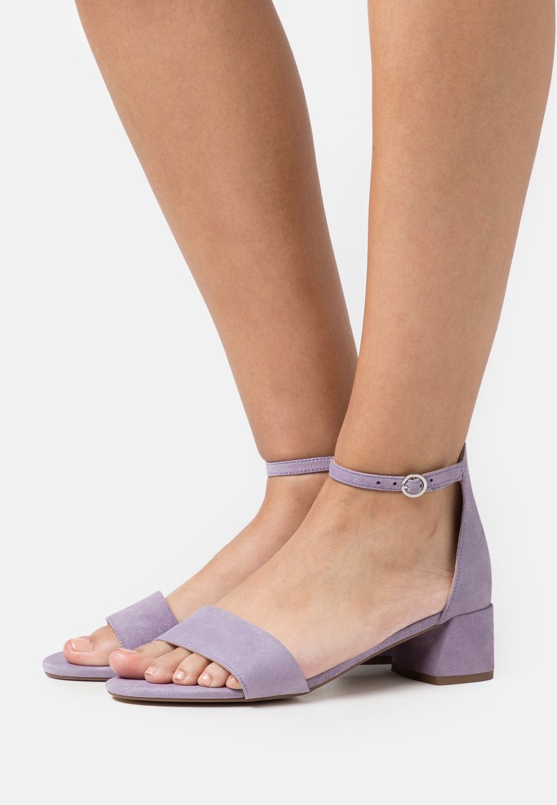 Pieds portant des sandales à bout ouvert violet clair avec bride à la cheville et petits talons blocs sur un fond uni clair.