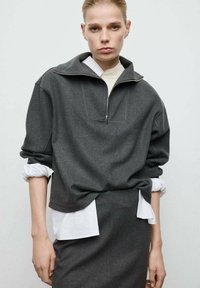 Gray zip-up sweatshirt with a wide collar, made of soft fabric. Layered over a white shirt, features long sleeves with rolled cuffs.