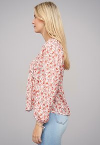 Blusa floreale a manica lunga in rosa pastello, caratterizzata da fiori arancioni e viola, con un fronte arricciato e una vestibilità rilassata. Tessuto leggero e texture morbida.