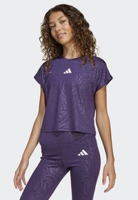 Young woman wearing purple animal-print sports top and leggings with white Adidas logos, standing against a plain light background.