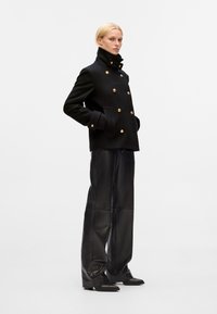 Zwarte wollen double-breasted jas met gouden knopen, hoge kraag en zijzakken. Gecombineerd met zwarte leren brede broek en elegante schoenen.