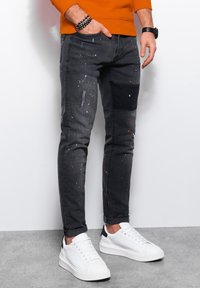 Mustat farkut, joissa on maalitahroja, suunniteltu kapealla leikkauksella, jossa on applikoitu osa, ja ne on yhdistetty valkoisiin lenkkareihin.