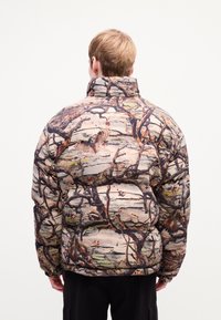 Person set fra bagfra iført en pufferjakke med et detaljeret skov camouflage mønster, der inkluderer grene, ugler og fugle.