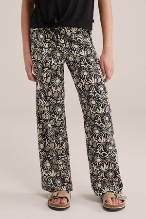 Zijaanzicht van een persoon die zwart-beige wijde broek met bloemenpatroon draagt en beige sandalen met open tenen en gespen.