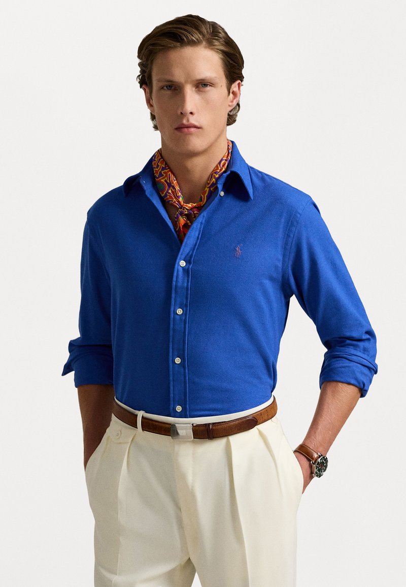 Blue button-up shirt with long sleeves, featuring a classic collar. Worn with a patterned orange scarf, cream trousers, and a brown belt.