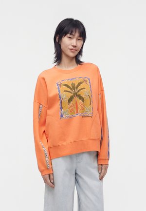 Ung kvinde iført en orange sweatshirt med et palmetræsgrafik og mønstrede ærmer, kombineret med lyseblå jeans, stående mod hvid baggrund.
