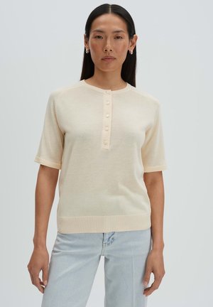 Vrouw met lang donker haar, gekleed in een crème kortärmelig vest met knopen en lichte blauwe spijkerbroek, staand tegen een effen lichte achtergrond.