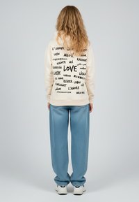 Felpa beige chiaro con testo nero in diverse lingue, in evidenza la parola "AMORE"; abbinata a pantaloni larghi blu chiaro e sneakers bianche.