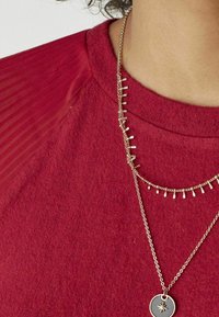 Rotes strukturierter Hemd mit Netzakzenten. Zwei goldene Halsketten: eine mit kleinen Anhängern, die andere mit einem runden Anhänger mit einem Sterndesign.