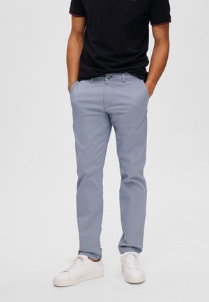 Lichtblauwe broek van een gladde stof, met voorzakken, een knoopsluiting en taps toelopende benen. Gekleed met witte sneakers.