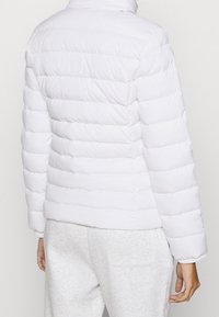Biała kurtka puchowa z poziomym przeszyciem, wysoka kołnierz i gładka tekstura, w połączeniu z szarymi dresami. Brak widocznych elementów metalowych.