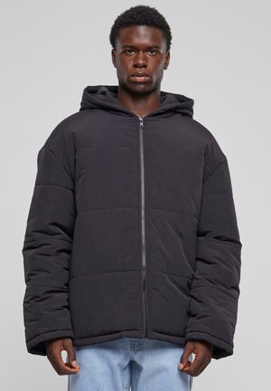 Chaqueta acolchada negra con capucha, que cuenta con un cierre frontal, forma cuadrada y tejido texturizado. Las costuras visibles crean un patrón acolchado.