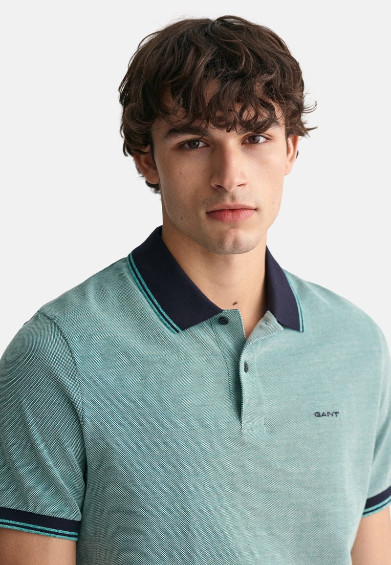 Türkises Poloshirt mit kurzen Ärmeln aus strukturiertem Stoff, marineblauem Kragen und türkisfarbenen Akzenten; mit Knopfleiste und kleinem Logo auf der Brust.