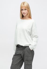 Világosszürke pulóver széles nyakkivágással és leeresztett vállakkal, sötétszürke cargo nadrággal párosítva, mely több zsebbel és varrási részletekkel rendelkezik.