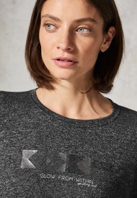 Tmavě šedé tričko s krátkým rukávem, které má texturovaný grafický potisk s šipkami a textem "ZÁŘÍ Z TOHO NEJEN MIMO" v bílé barvě. Hladký povrch látky.