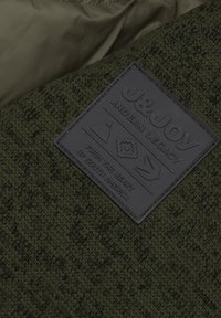 Gestreepte donkergroene stof met een rechthoekige zwarte leren patch met verhoogde ontwerpen en tekst, die het merk en de oorsprong aangeeft.