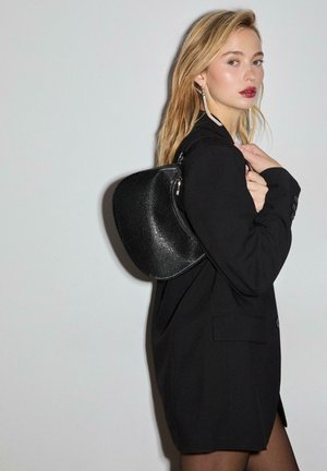 Borsa nera con una finitura lucida e testurizzata, forma curva, dettagli in argento, indossata a spalla, abbinata a un blazer nero.
