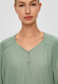 Lichte groene blouse met een V-hals, korte knoopsluiting en geplooide schouders. Gemaakt van een gestructureerde, lichte stof.