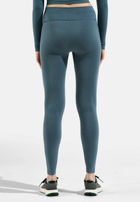 Gerippte, hochtaillierte Leggings in dunklem Blaugrün mit einer figurbetonten Passform und knöchellanger Schnittführung, kombiniert mit schwarzen und grauen Sportschuhen.