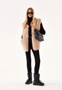 Gilet in pelliccia sintetica rosa chiaro, dolcevita nero, jeans skinny neri e stivali neri trapuntati. Grande borsa trapuntata con tracolla a catena. Occhiali da sole neri.