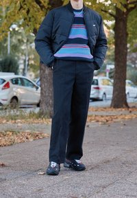 Vadderad svart bomberjacka, färgglad randig stickad tröja, mörka byxor och svarta sneakers med röda accenter. Står på en trottoar.