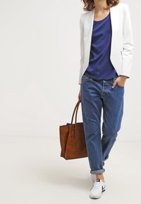 Blazer bianco su misura sopra una maglia di raso blu navy, abbinata a jeans blu risvoltati e sneakers bianche. Tiene una borsa tote in pelle marrone.