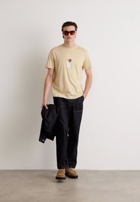 Beige bomulds t-shirt med en lille grafik, sorte denim bukser, iført brune sko og solbriller, holder en sort jakke. Enkel, moderne stil.